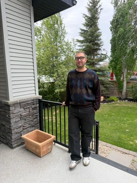 Man wearing glasses and an argyle sweater stands on a deck overlooking a green lawn.